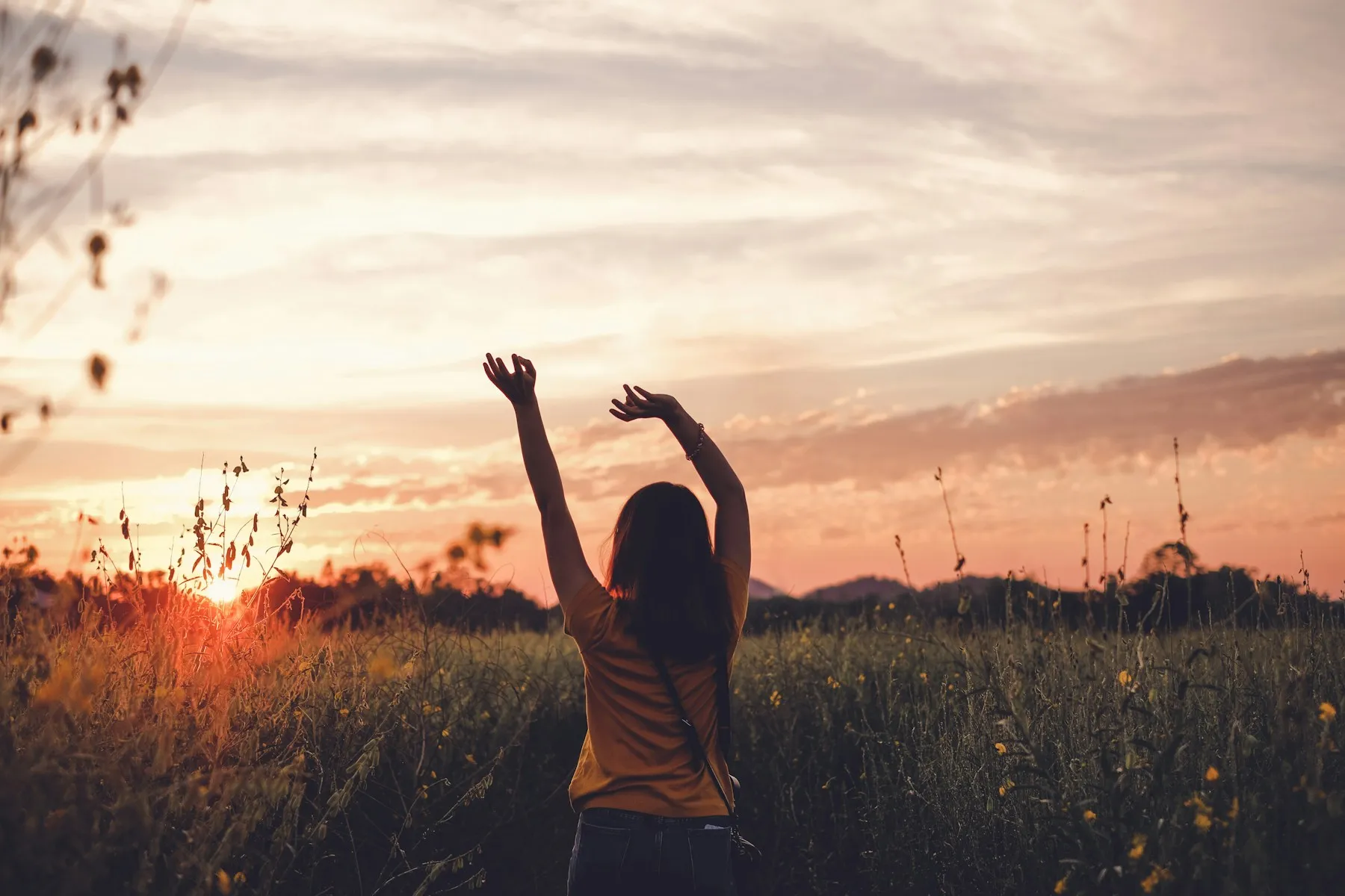 A woman in a wildflower meadow raising both arms toward a glowing sunset sky