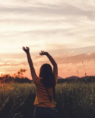 A woman in a wildflower meadow raising both arms toward a glowing sunset sky