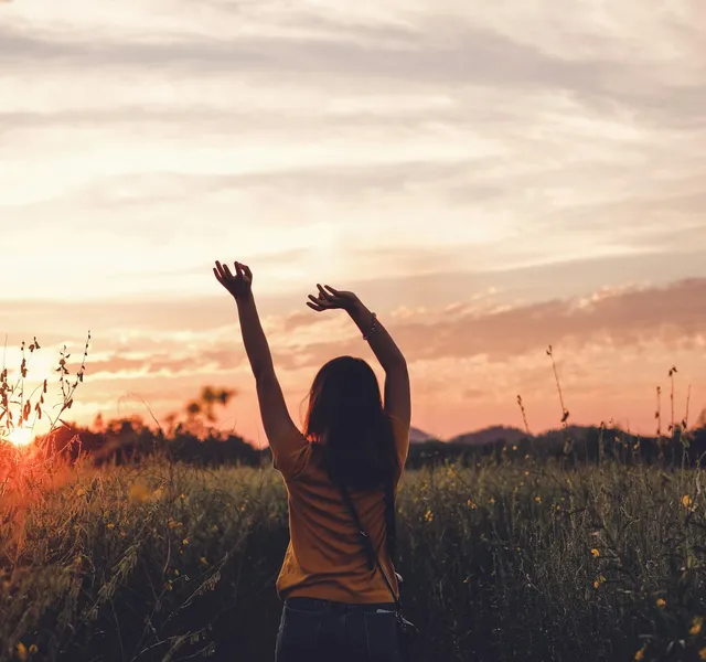 A woman in a wildflower meadow raising both arms toward a glowing sunset sky