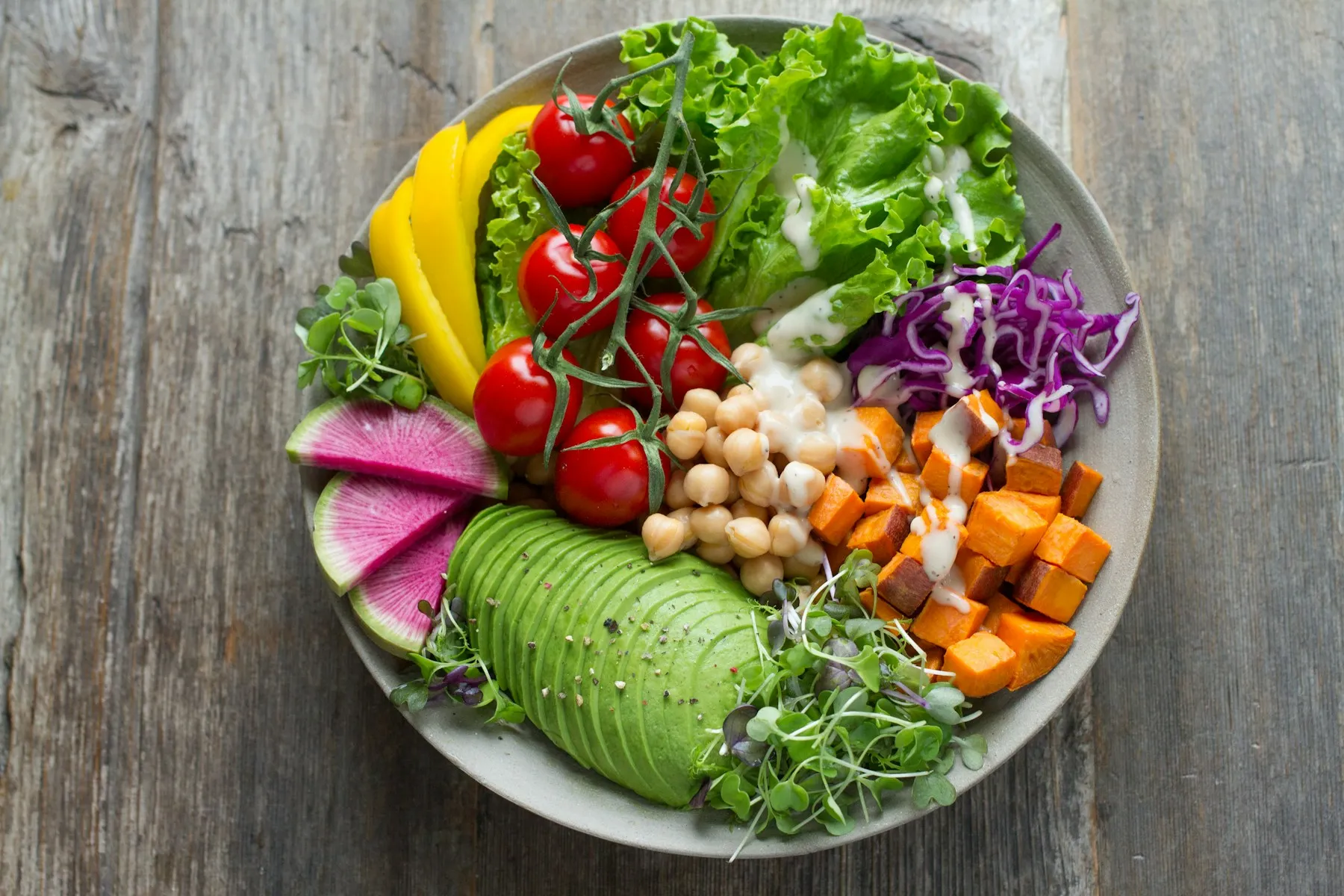 A bowl piled with avocado, vine tomatoes, chickpeas, sweet potato, radish, and red cabbage on a wooden table