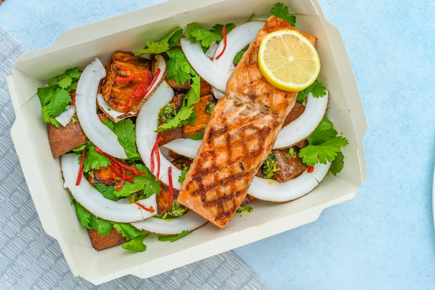 A grilled salmon fillet with lemon over roasted sweet potato, coconut, and cilantro in a takeaway box