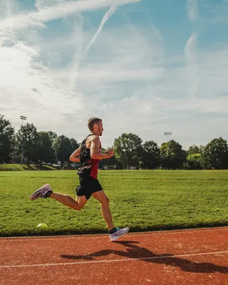A runner mid-stride on a red running track beside a sunlit green field under a blue sky