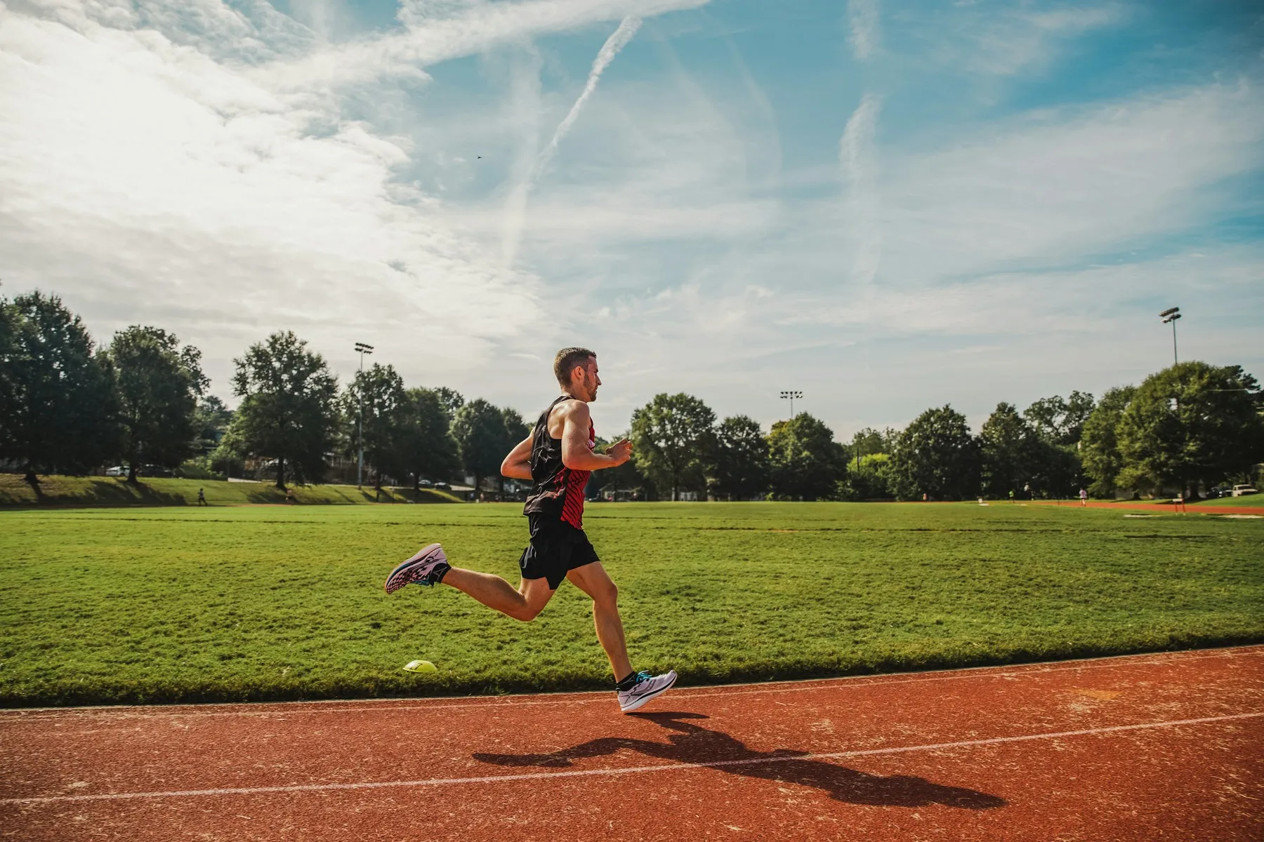 A runner mid-stride on a red running track beside a sunlit green field under a blue sky